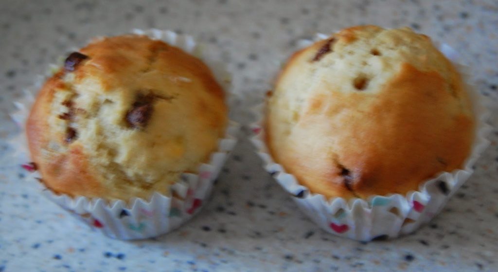Two golden-brown banana chocolate chip muffins sit side by side in white paper cases on a speckled worktop.