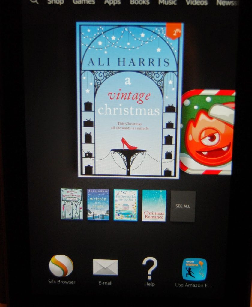 Tablet screen displaying an ebook library on a Kindle Fire. The highlighted book is "A Vintage Christmas" by Ali Harris, featuring a snowy cover with presents and a woman in red. Various other book covers and app icons are visible below.