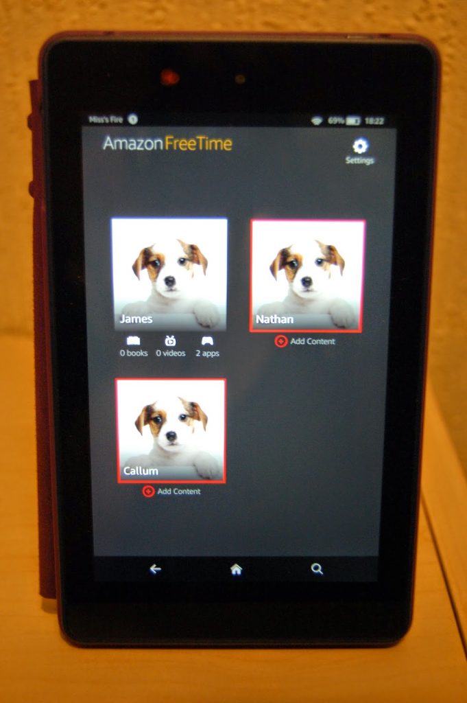 A Kindle Fire HD 6 tablet screen displays the Amazon FreeTime app with three user profiles, each showing a puppy image and labelled James, Nathan, and Callum. Two profiles display "Add Content." The device rests on a wooden surface.