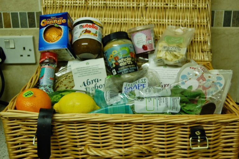 A wicker basket filled with assorted food items—including fruit, Nutella, peanut butter, chocolate, dried pasta, spinach, juice, seasonings, and Abra-ca-Debora pancakes—sits on a kitchen worktop, perfect for Pancake Day celebrations.