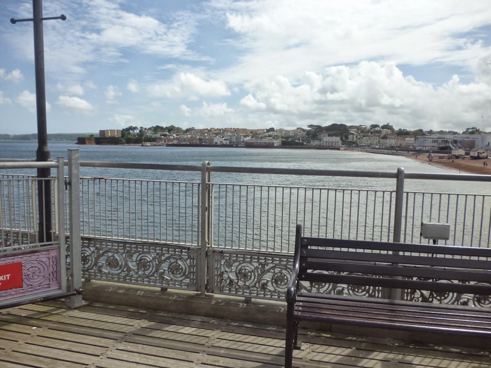 A wooden pier with a metal railing overlooks calm water and a coastal town in the distance. For this Flashback Friday, an empty black bench sits quietly in the foreground under a partly cloudy sky.
