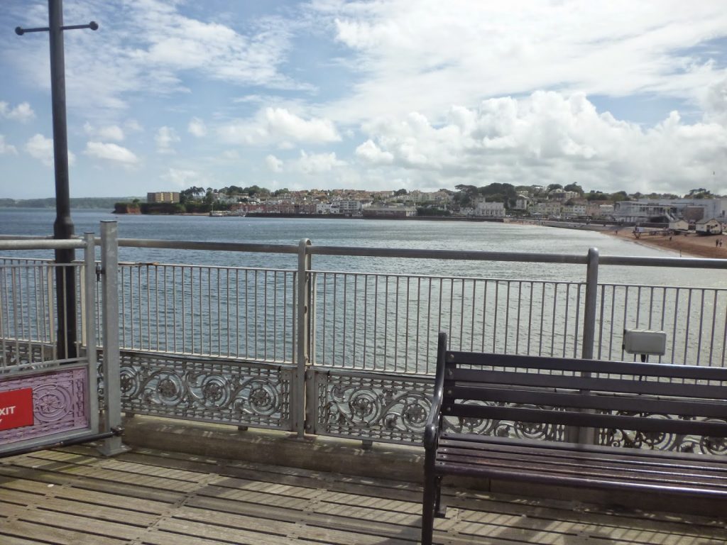 A wooden pier with a metal railing overlooks calm water, with a coastal town and sandy beach visible in the distance under a partly cloudy sky. An empty bench sits in the foreground, perfect for a quiet Flashback Friday moment.
