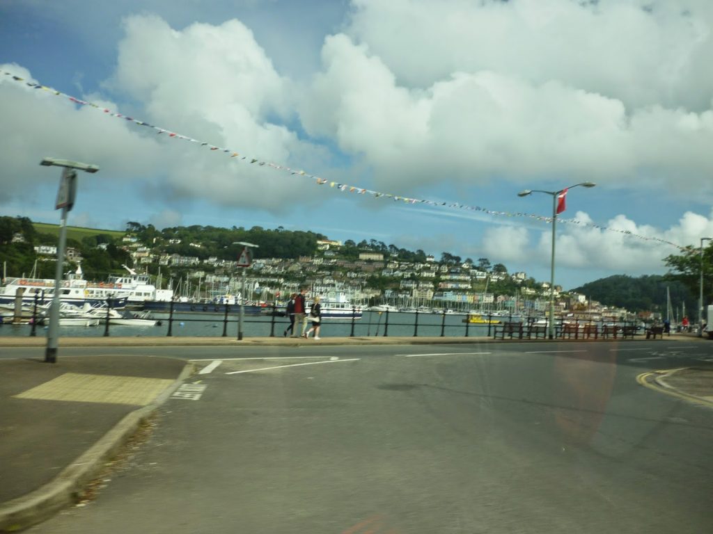 A scenic view of a harbour with boats moored, houses on a green hillside, bunting overhead, and a few people strolling along the waterfront under a partly cloudy sky for a perfect Flashback Friday memory.