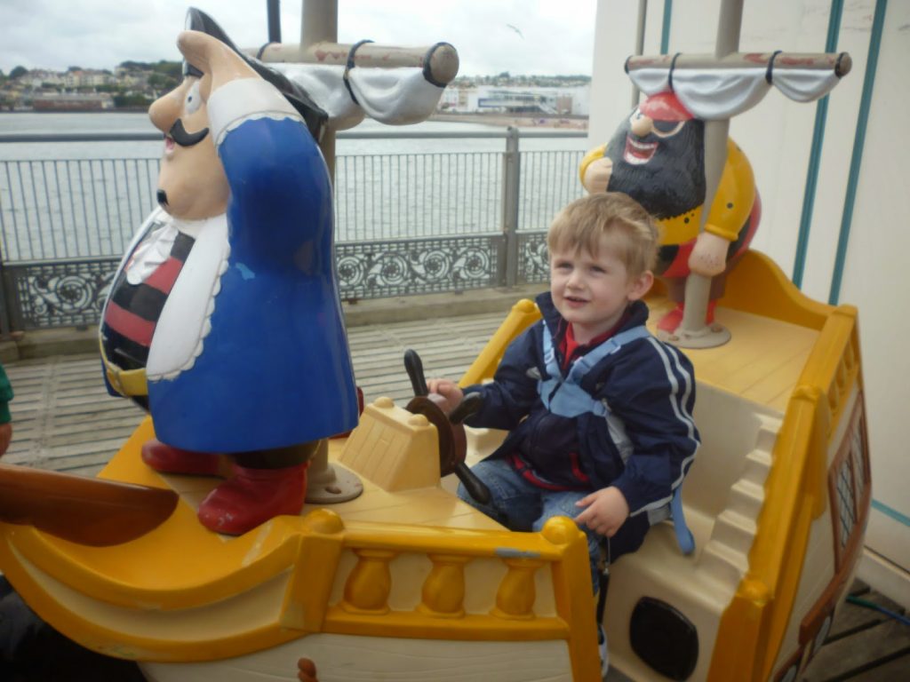 A young boy sits on a coin-operated pirate ship ride, smiling and holding the steering wheel, with colourful pirate statues beside him. This joyful Flashback Friday moment is set on a lively promenade near the water.