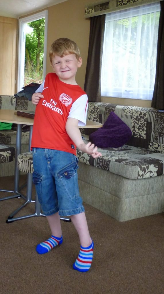 Flashback Friday: A young boy in an Arsenal football shirt, denim shorts, and colourful socks pretends to play air guitar with a purple cushion in a cosy living room with patterned sofas and large windows.