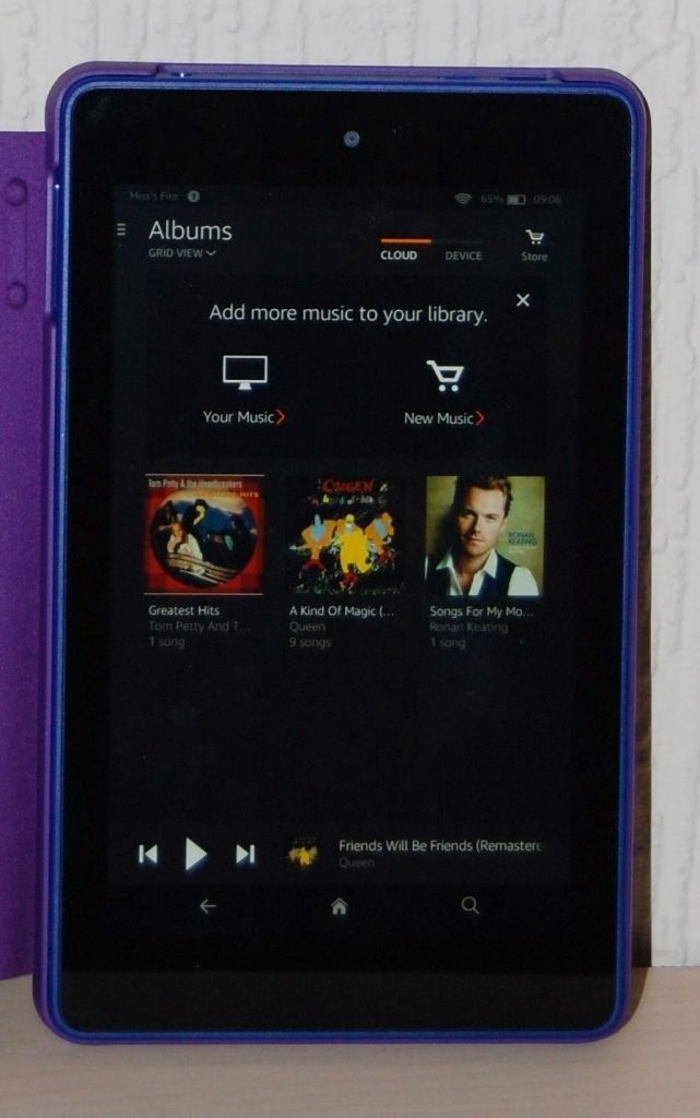 A Kindle Fire HD 6 displays a vibrant music library screen with album covers like Greatest Hits by Queen and Songs for My Mother by Ronan Keating. This tablet review highlights the easy interface prompting users to add more music.