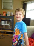 Flashback Friday: A young boy with fair hair, wearing a blue T-shirt with colourful cartoon characters, stands and smiles in a cosy living room with a sofa, window, and fireplace in the background.