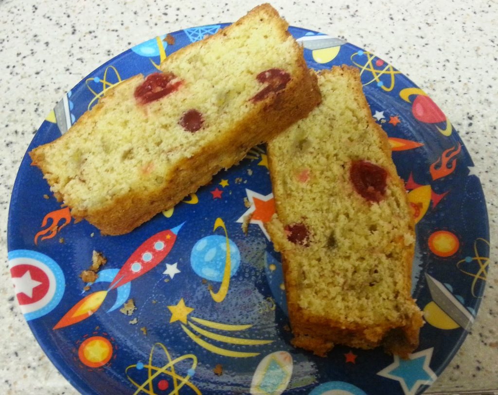 Two slices of fruit cake with visible red glacé cherries sit on a colourful plate decorated with rockets, planets, and stars—perfect for a peaceful Silent Sunday on the speckled worktop.
