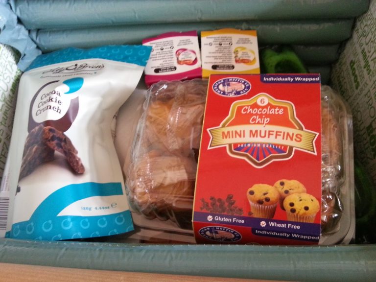 A snack subscription box featuring a bag of cocoa biscuit crunch, six chocolate chip mini muffins, and three condiment sachets, all packed together in green insulated packaging.