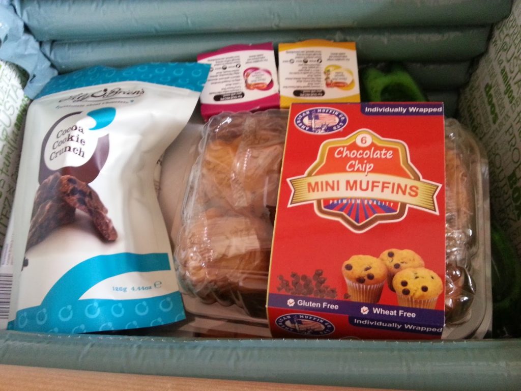 A Degustabox contains a bag of cocoa cookie crunch, gluten and wheat free chocolate chip mini muffins, and condiment sachets, all arranged in green bubble wrap packaging—perfect for your next snack subscription discovery!.