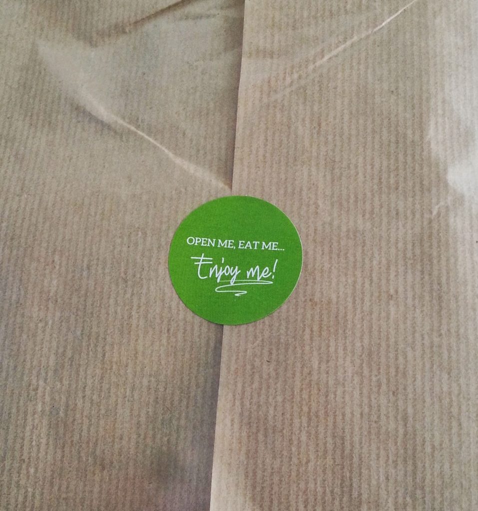 A brown paper parcel sealed with a round green sticker that reads, "OPEN ME, EAT ME... Enjoy me!" in white text—perfect for your March Degustabox snack box review.