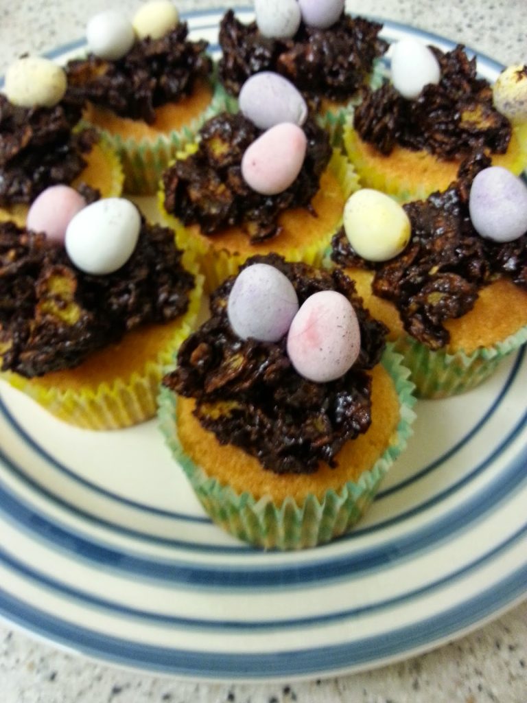 Easter cupcakes topped with nests of chocolate-coated cereal and pastel-coloured sweet eggs are arranged on a blue-and-white striped plate, making delightful Easter desserts.