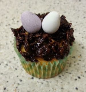 An Easter Nest Cupcake with chocolate topping designed to look like a nest, topped with two egg-shaped sweets—one purple and one white—rests on a speckled worktop.