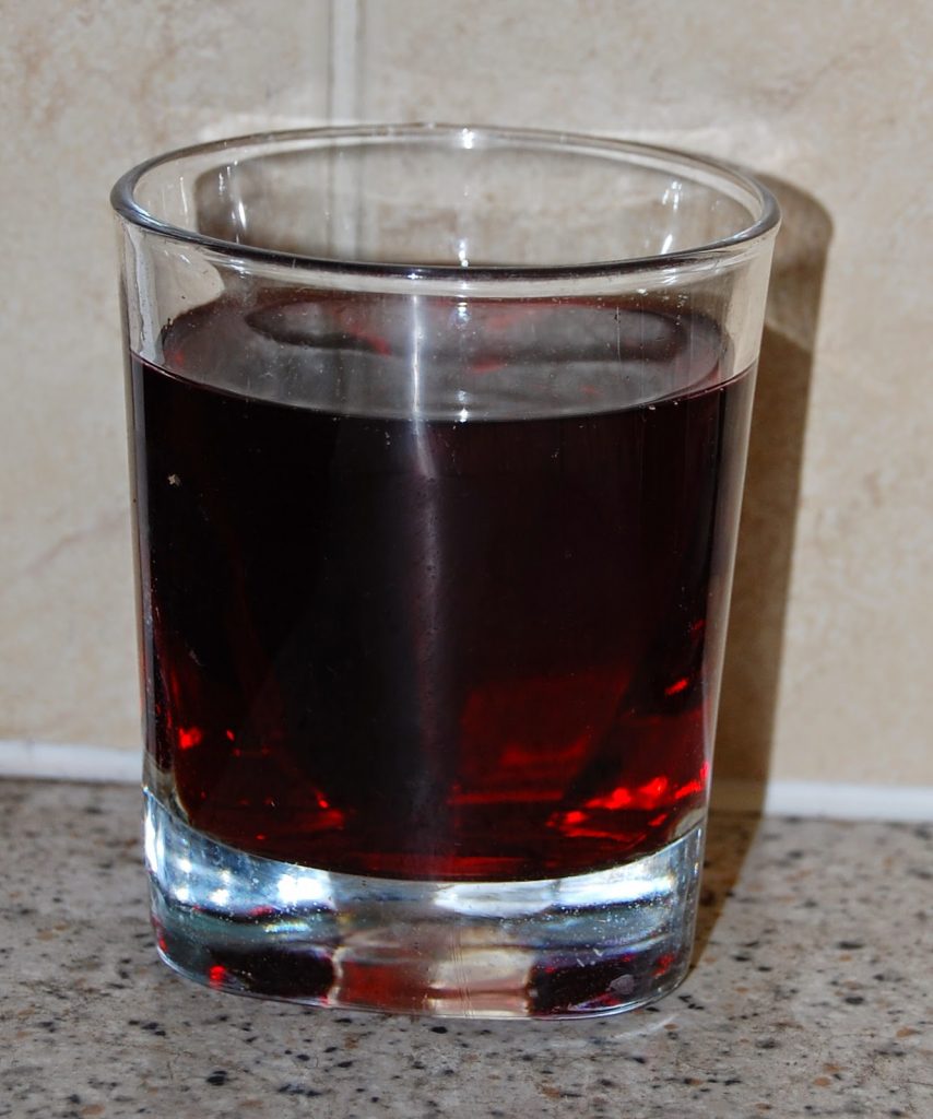 A clear glass filled with dark red cherry juice sits on a speckled worktop, with a beige tiled wall in the background—perfect to start your day with some good juice.