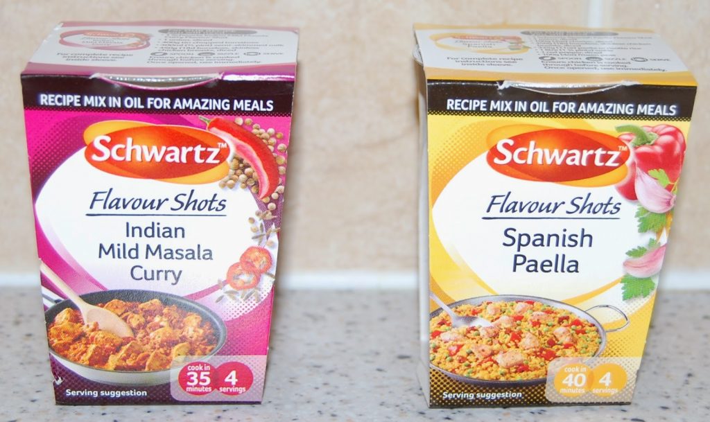Two boxes of Schwartz Flavour Shots sit on a kitchen worktop—Indian Mild Masala Curry and Spanish Paella—each displaying meal images, serving suggestions, and cooking times, perfect for a food box review or snack subscription box feature.