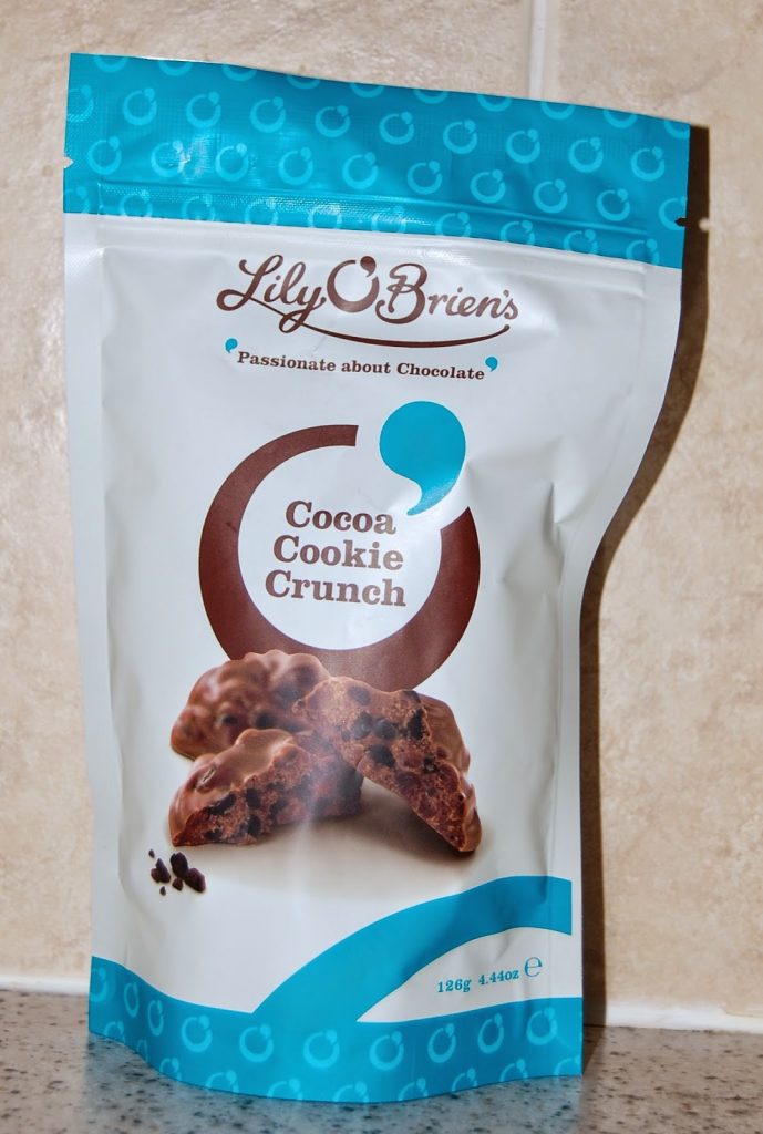 A standing pouch of Lily O'Brien's Cocoa Cookie Crunch chocolates with an image of chocolate biscuits on the front, featured in a recent Degustabox review, sits on a speckled worktop against a tiled wall background.
