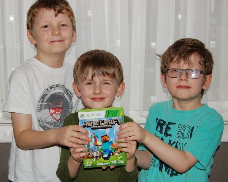 Three young boys stand together indoors, smiling and holding an Xbox 360 Minecraft game case. Two wear glasses as they celebrate with friends, learning about the value of money through saving up for their favourite game.