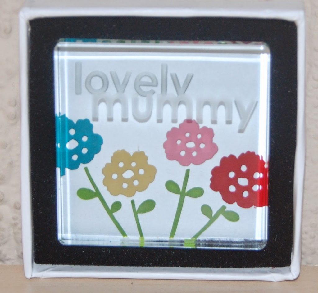 A glass plaque in a black and white box, featuring colourful flowers and the words "lovely mummy" engraved above them—a thoughtful addition to any Pamper Hamper.