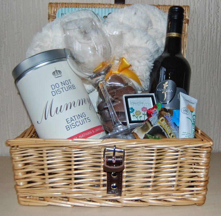 This Pamper Hamper features a wicker basket with a wine bottle, wine glass, “Do Not Disturb Mummy Eating Biscuits” tin, biscuits, snacks, fluffy blanket, and bath products—all secured with a leather strap.