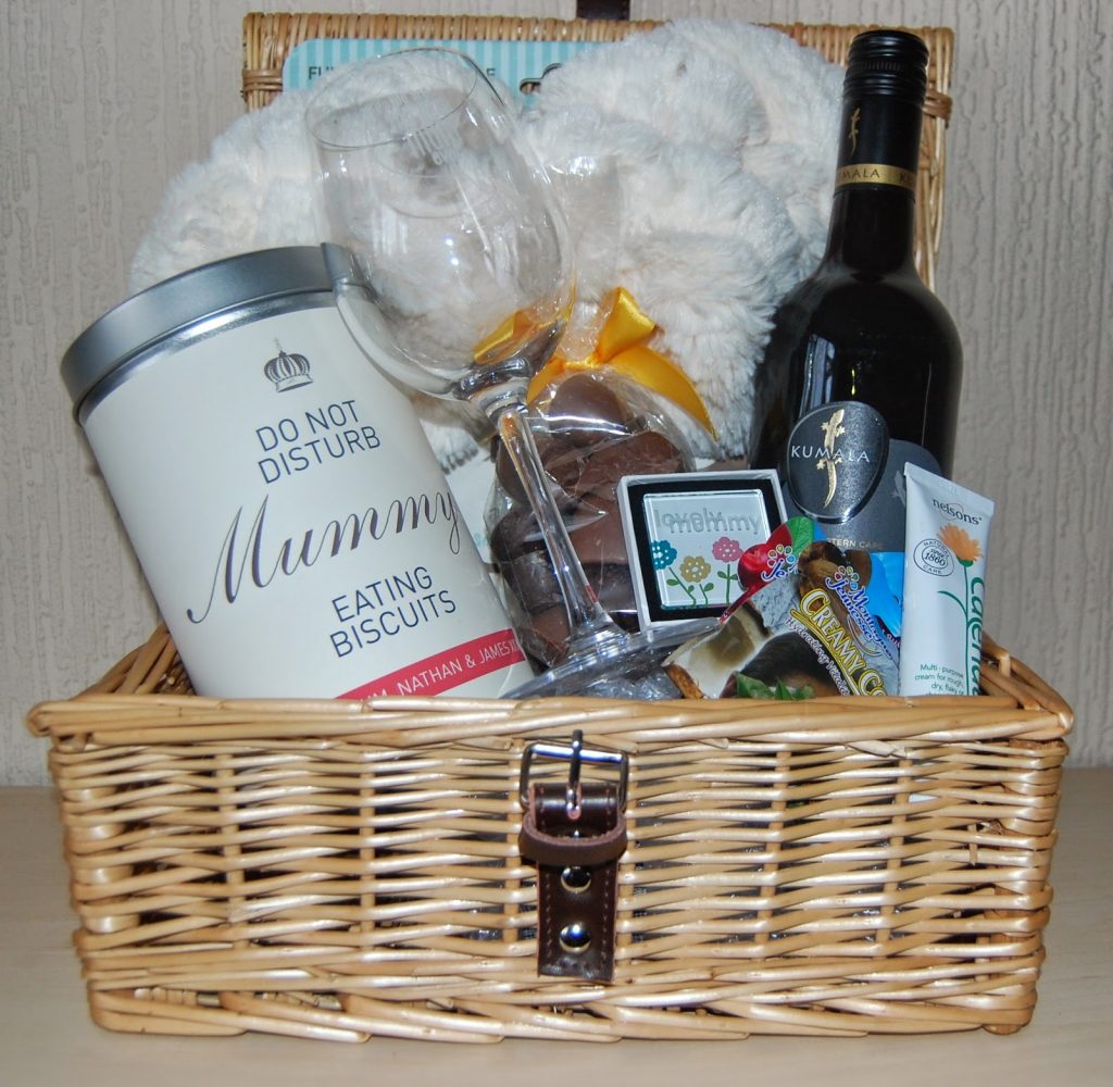A Pamper Hamper wicker gift basket contains a bottle of red wine, a tin labelled "Do Not Disturb Mummy Eating Biscuits," biscuits, chocolates, a wine glass, a fluffy blanket, and various small treats.
