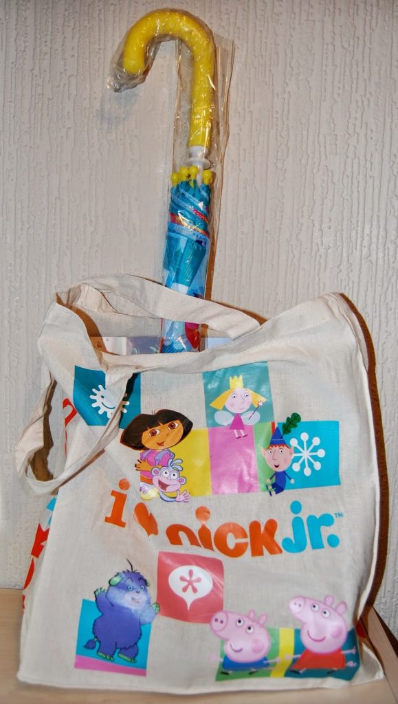 A white tote bag with colourful Nick Jr. characters, including Dora the Explorer, Peppa Pig, and Blaze and the Monster Machines, stands upright with a yellow-handled umbrella in plastic sticking out. The bag is against a textured wall—perfect for your next Twitter party!.