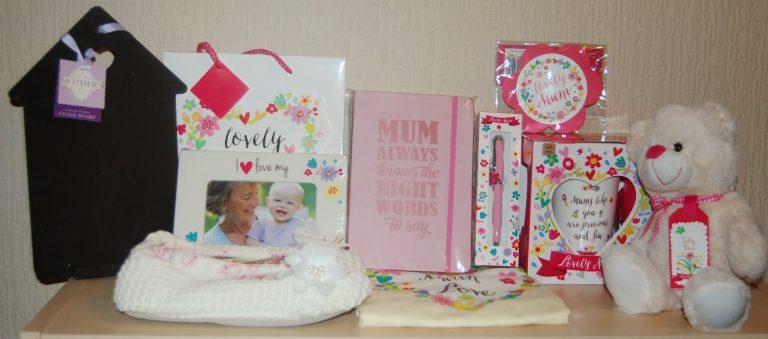 A collection of Mother’s Day gifts from Home Bargains sits on a shelf, including a white teddy bear, floral-themed mugs and stationery, a knitted hat, framed photos, a decorative plaque, and a pink notebook that reads "Mum always knows the right words to say.
