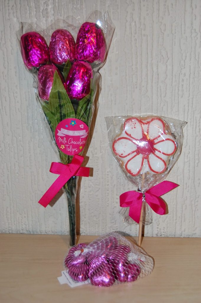 A bouquet of pink foil-wrapped chocolate tulips, a flower-shaped biscuit on a stick with white icing, and a mesh bag of pink foil-wrapped chocolate hearts—all tied with pink ribbons—make charming Mother’s Day gifts from Home Bargains, displayed against a light textured wall.