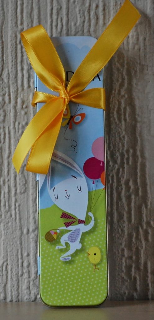 A rectangular tin box decorated with a cartoon rabbit and chick, balloons, and a butterfly is tied with a bright yellow ribbon and bow—an adorable choice for Alternative Easter Gifts—standing upright against a textured beige background.