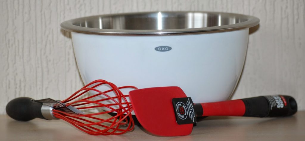A large white mixing bowl with a stainless steel rim sits on a worktop. In front of the bowl are tools perfect for making Easter Nest Cupcakes—a red silicone whisk and a red silicone spatula, both with black handles.