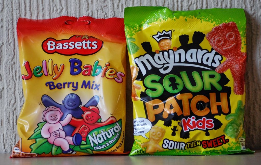 Part of the March Degustabox Review, two colourful sweet bags: one for Bassetts Jelly Babies Berry Mix with cartoon jelly baby characters, and one for Maynards Sour Patch Kids featuring illustrated sour sweets on a green and yellow background.