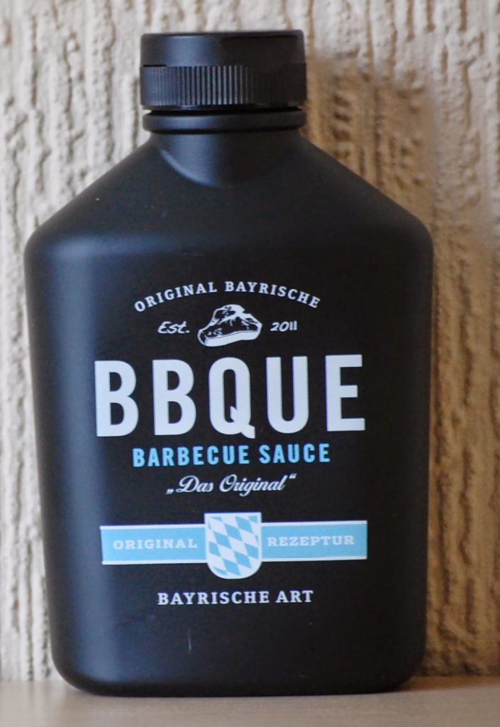 A black bottle labelled “BBQUE Barbecue Sauce – Das Original,” featuring a Bavarian-themed design and blue and white chequered shield, as seen in the March Degustabox review, placed against a textured beige background.