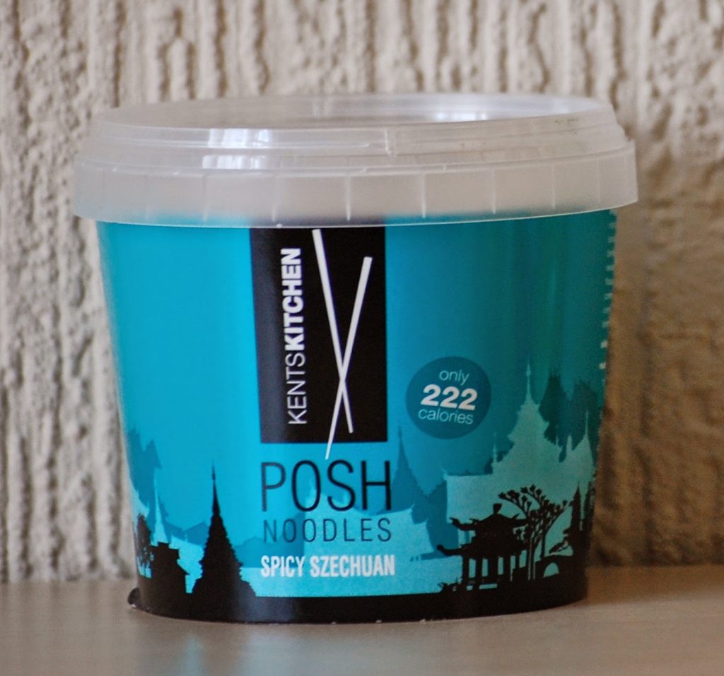 A blue container of Kent’s Kitchen Posh Noodles, Spicy Szechuan flavour, sits on a wooden surface. Featured in the March Degustabox Review, the packaging notes "only 222 calories" and showcases an Asian-inspired skyline design.