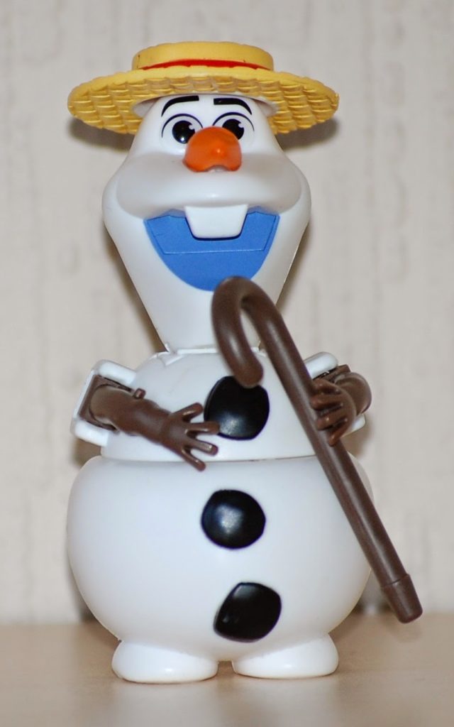 A toy snowman with a big blue smile, orange carrot nose, black buttons, stick arms, and a yellow straw hat holds a brown walking stick against a light patterned background—perfect as alternative Easter presents.