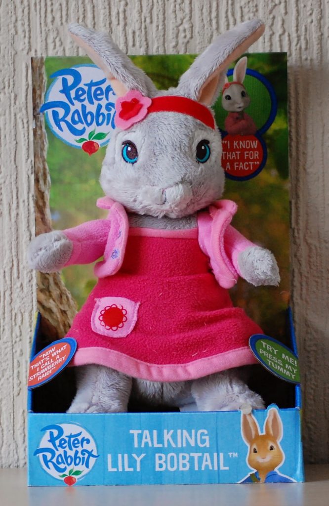 A plush Talking Lily Bobtail toy from Peter Rabbit is displayed in a blue and green box. Wearing a pink dress and flower headband, this adorable toy makes sounds when pressed—perfect as an Alternative Easter Gift.