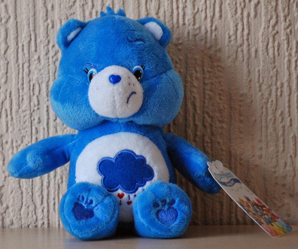 A blue plush bear with a dark blue cloud and heart symbols on its belly sits upright against a textured beige wall. With its slightly frowning expression, this bear makes a unique choice for Alternative Easter Gifts. Tag attached to its left side.