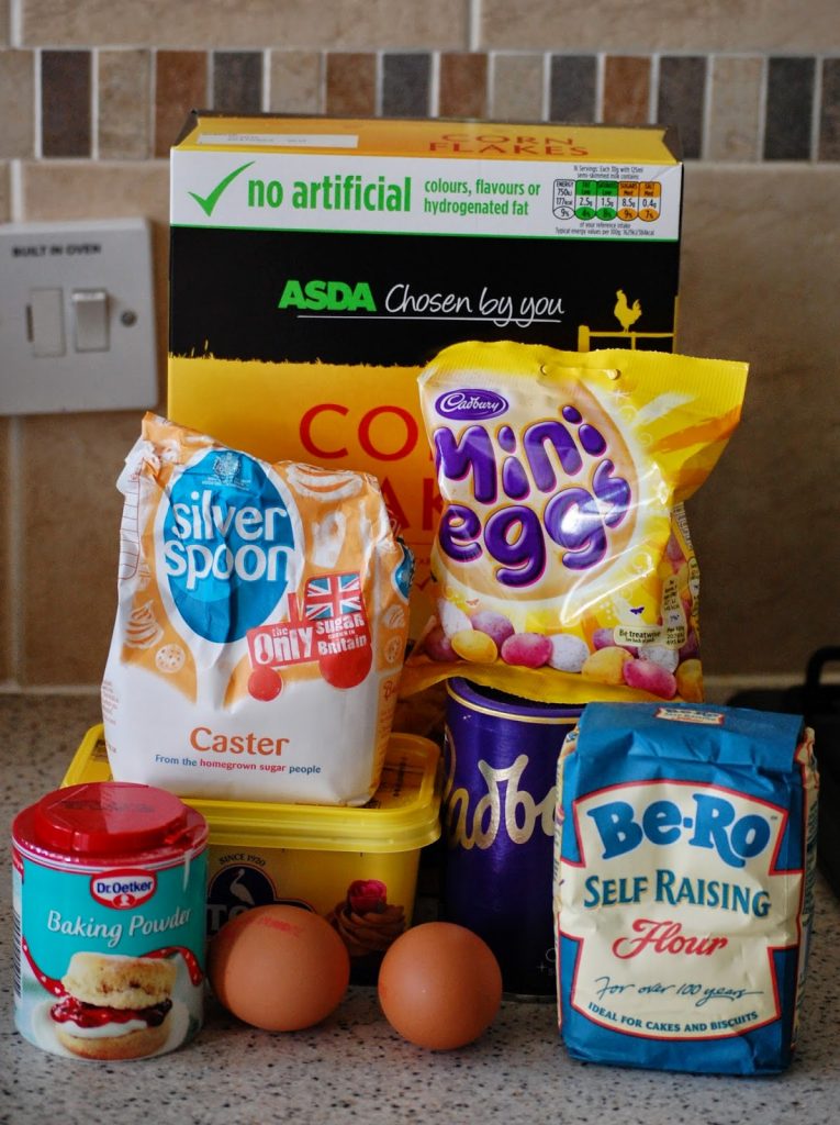 A variety of baking ingredients on a kitchen worktop, perfect for making Easter desserts like nest cupcakes, including caster sugar, baking powder, self-raising flour, margarine, cornflakes, mini eggs, two eggs, and a mixing bowl.