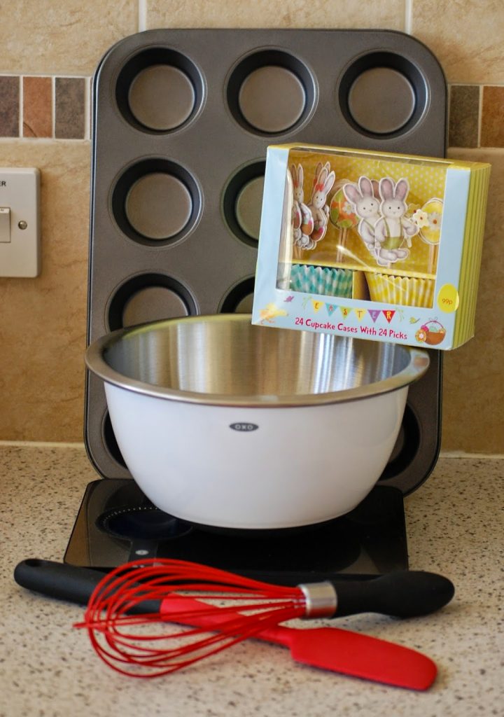 A mixing bowl, muffin tray, cupcake cases with bunny picks, red whisk, and black spatula are arranged on a kitchen worktop—perfect for making Easter Nest Cupcakes.