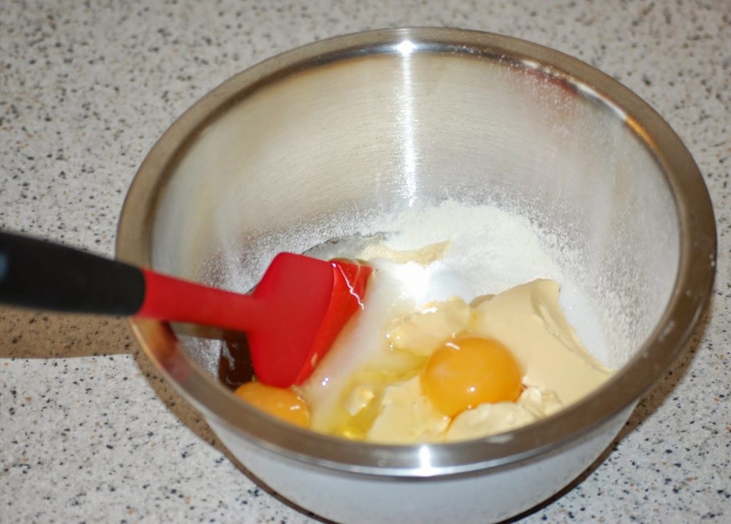 A metal mixing bowl on a speckled worktop holds the ingredients for Easter Nest Cupcakes—eggs, flour, sugar, and butter. A red spatula rests inside the bowl, ready to mix everything together.
