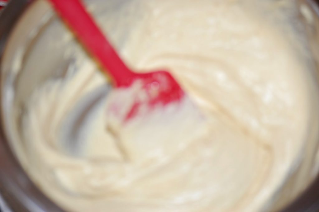A red spatula stirs a creamy, light-coloured batter in a mixing bowl, perhaps for delicious Easter Nest Cupcakes. The image is blurry and out of focus.