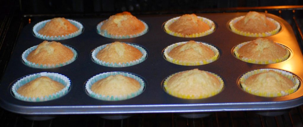 A muffin tin with twelve freshly baked Easter Nest Cupcakes in colourful paper cases, resting inside an oven. The golden-brown cupcakes are slightly domed on top, perfect for a festive treat.