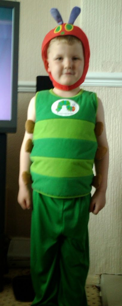 Flashback Friday: A young child is dressed in a green caterpillar costume with a red hood and purple antennae, inspired by "The Very Hungry Caterpillar." The child stands indoors, smiling at the camera.
