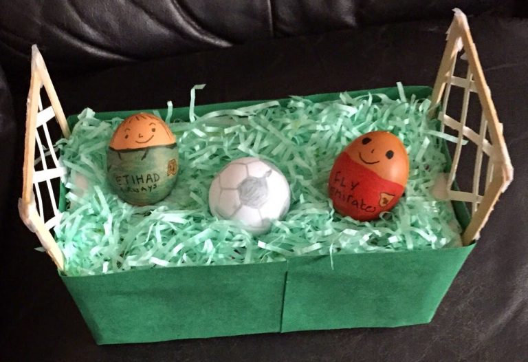 Two decorated eggs with smiling faces wear football shirts and stand in a green box resembling a football pitch, ready for a Silent Sunday match, with a toy football and paper goals at each end, surrounded by green shredded paper.