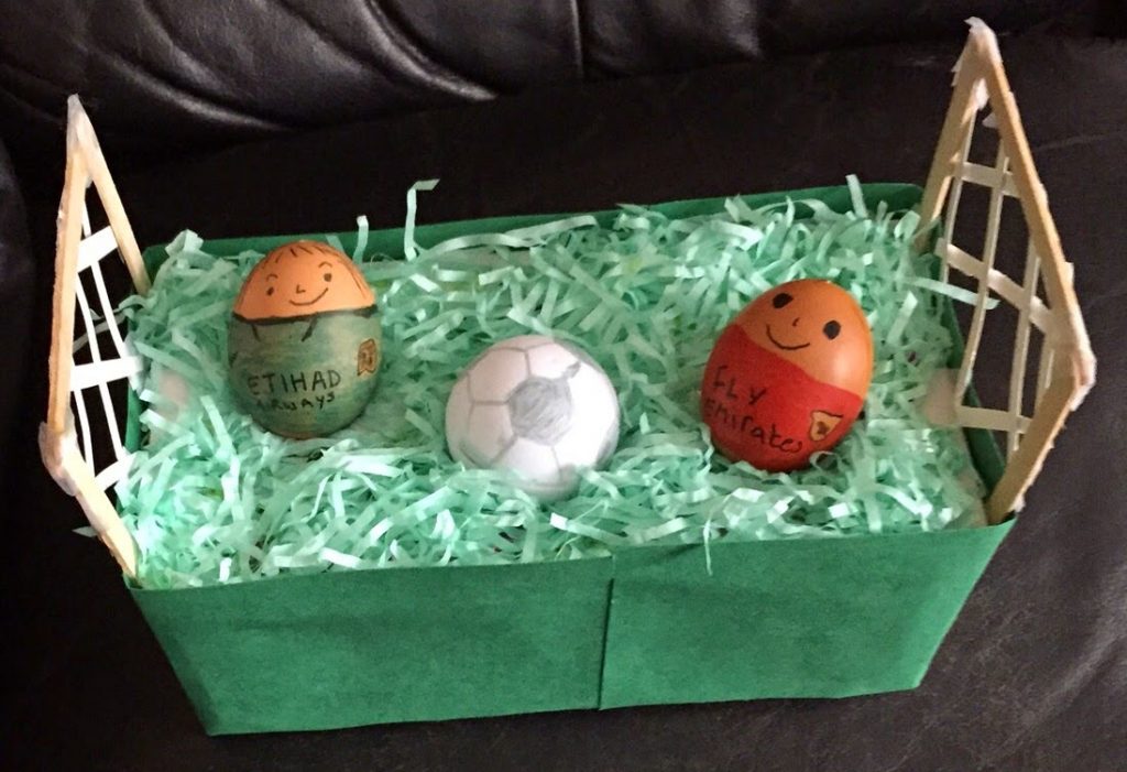 Two decorated eggs with smiling faces, dressed as footballers, are placed in a green box resembling a football pitch with paper grass and goalposts—a playful Silent Sunday scene complete with a toy football between them.
