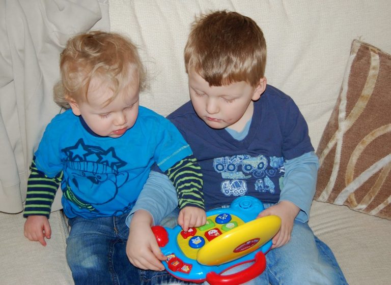 Flashback Friday: Two young children sit on a sofa, focused on playing with a colourful toy laptop. The older child helps the younger one press buttons, both dressed in blue shirts and jeans. A striped cushion is beside them.