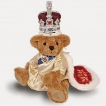 A regal teddy bear dressed in royal attire, complete with a crown and a cream-coloured cape with red lining and gold embroidery, sits upright against a white background.
