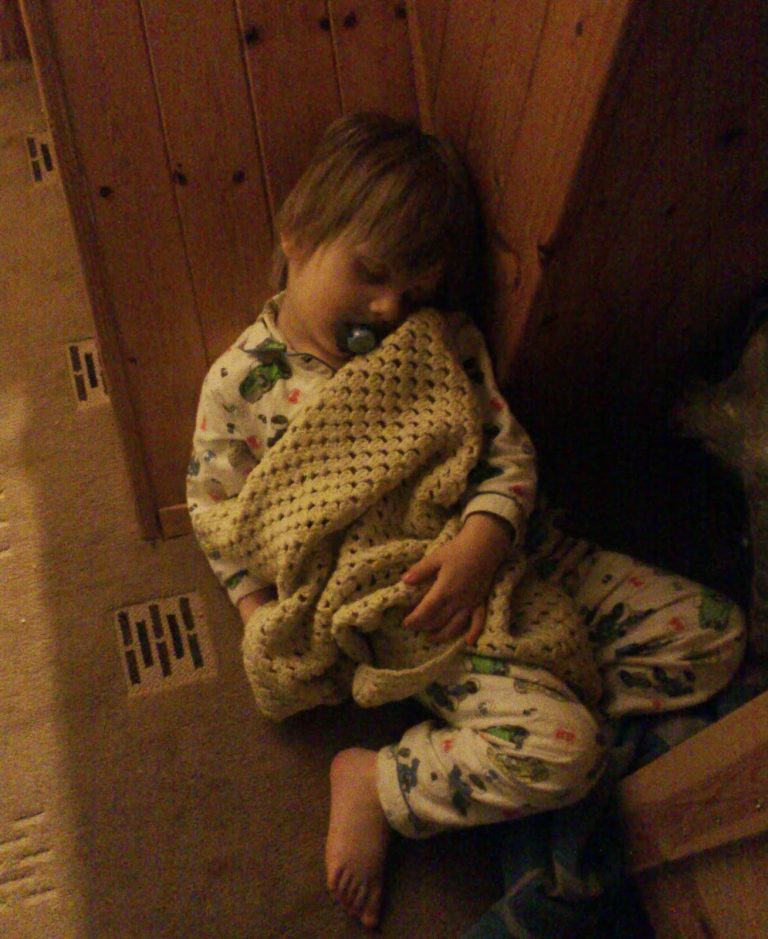 A young child in pyjamas sleeps upright against a wooden wall, holding a knitted blanket and using a dummy—an adorable Flashback Friday moment. The child’s head is tilted to the side, one bare foot is visible, and the lighting is dim and warm.