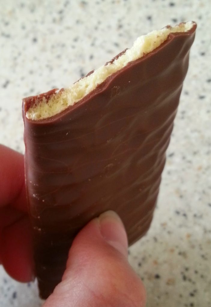 A hand holding a partially eaten rectangular chocolate-covered biscuit with a light, crumbly interior—one of those tasty little numbers—against a speckled worktop background.
