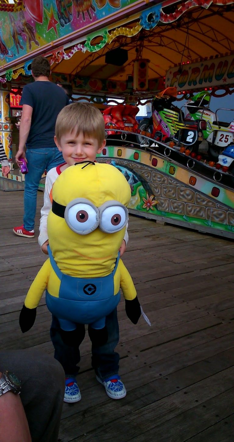 Flashback Friday: A young boy smiles whilst holding a large Minion plush toy at an amusement park, standing on a wooden boardwalk with colourful funfair rides and lights in the background.