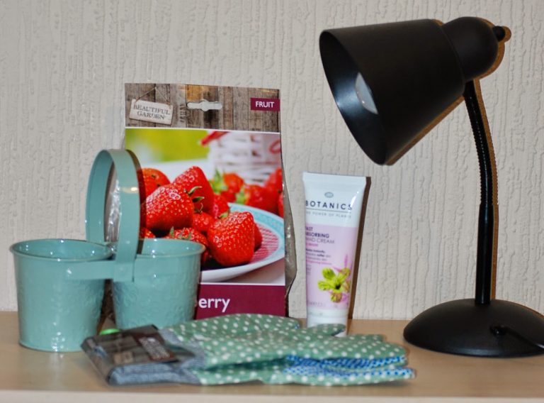 A black desk lamp, mint green plant pots, strawberry seed packet, tube of hand cream, green polka dot gardening gloves, and small gardening tools create a charming indoor garden scene on a light surface against a textured wall.