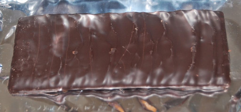 A rectangular chocolate-covered wafer bar rests on a crinkled metallic wrapper. This tasty little number features a dark, glossy coating with visible wavy lines on its surface.
